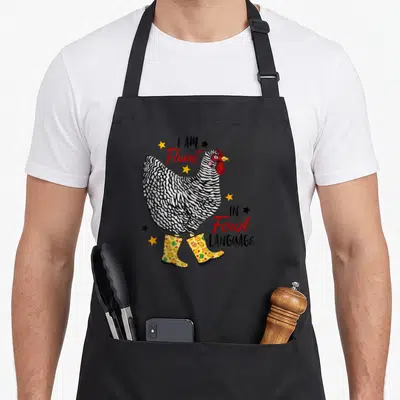 Fluent chicken with apron - Cartoon chicken in yellow boots with text I AM Fluent IN Fowl LANGUAGE and scattered stars — shown on a black cotton canvas apron against a white background