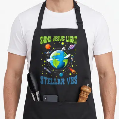 Stellar vacation bible school apron - Cartoon Earth with orbiting satellites, stars, and SHINE JESUS' LIGHT text — shown on a black cotton canvas apron against a white background