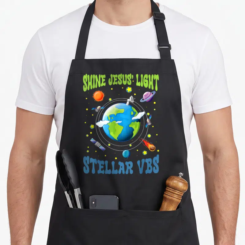Stellar vacation bible school apron - Cartoon Earth with orbiting satellites, stars, and SHINE JESUS' LIGHT text — shown on a black cotton canvas apron against a white background