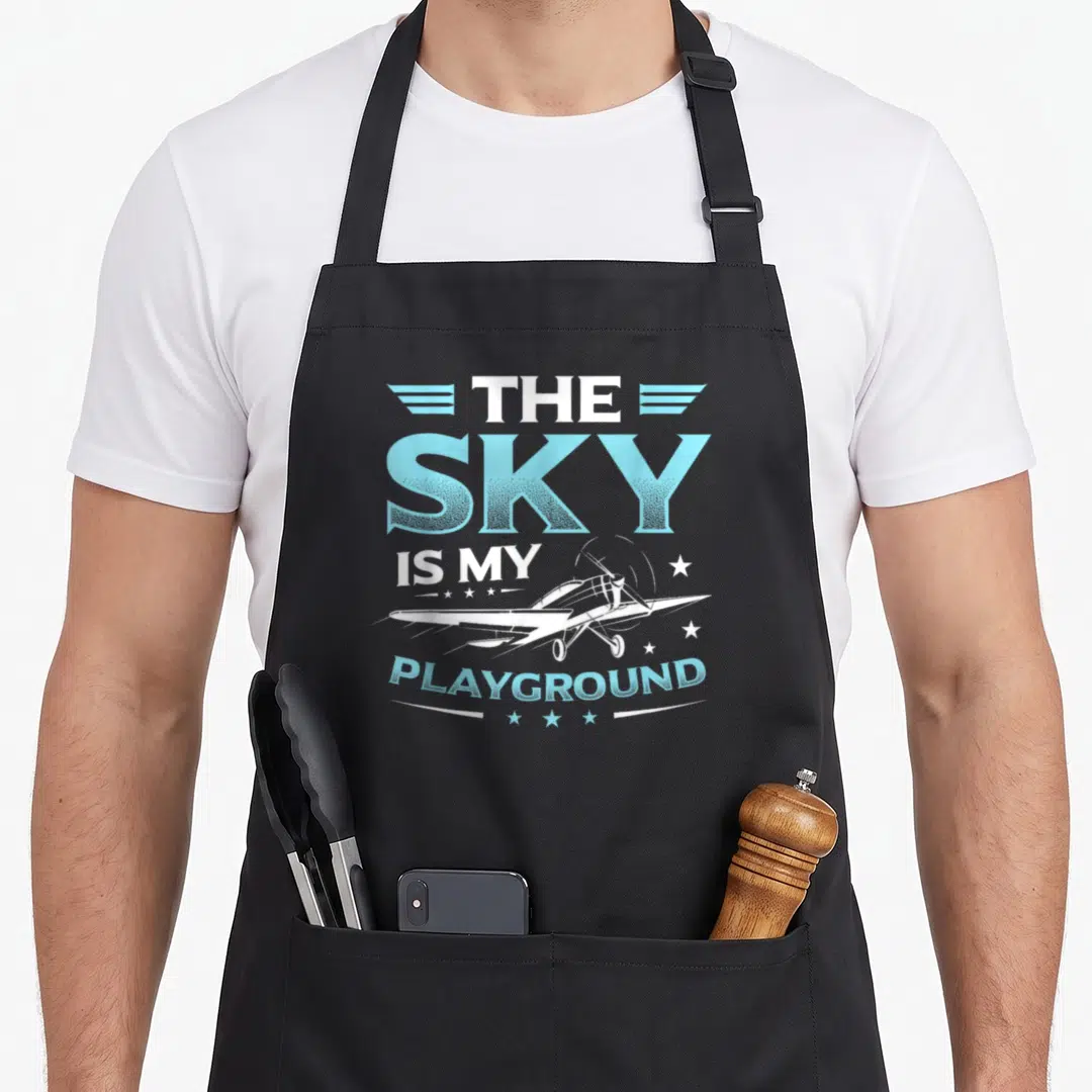 Sky airplane apron - The Sky Is My Playground text with white airplane and blue wings — shown on a black cotton canvas apron against a white background