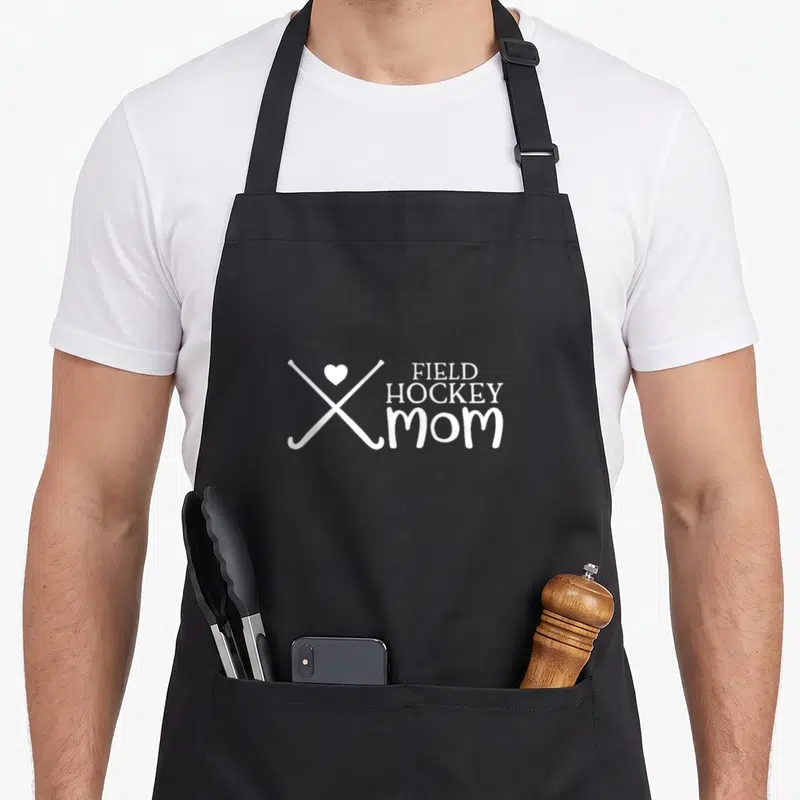 Field hockey mom apron - Two white crossed field hockey sticks with heart and stacked FIELD HOCKEY mom text — shown on a black cotton canvas apron against a white background