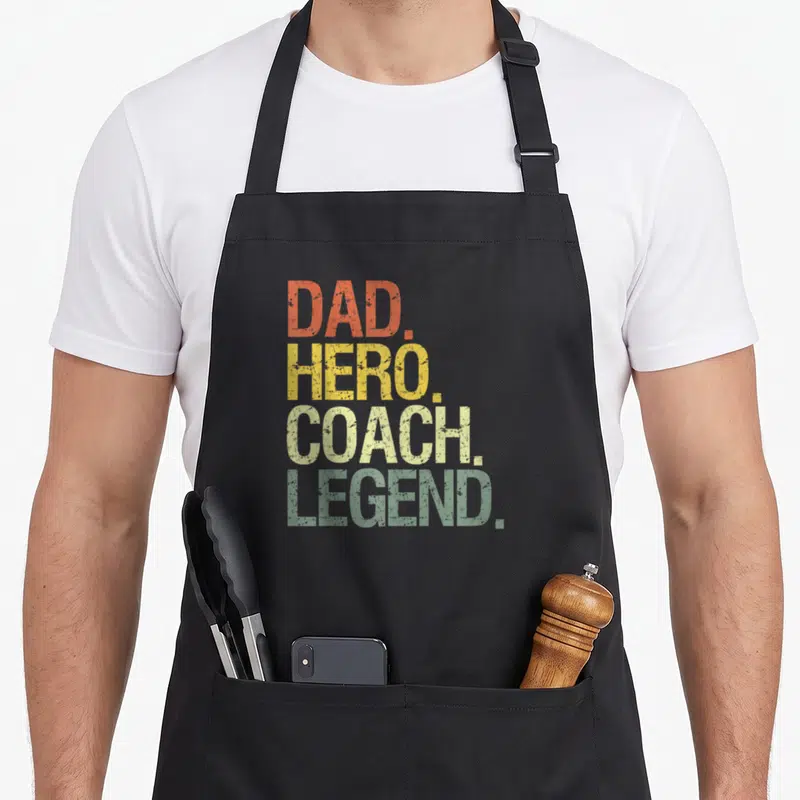 Funny coach sports legendz apron - Stacked DAD. HERO. COACH. LEGEND. text in orange, yellow, green and pale distressed — shown on a black cotton canvas apron against a white background
