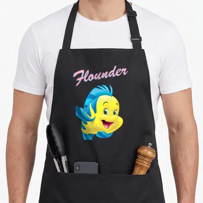 Wave mermaid apron - Pink Flounder text above smiling yellow fish with blue fins — shown on a black cotton canvas apron against a white background