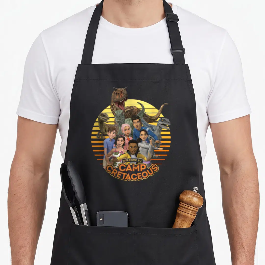 Camp jurassic world apron - Animated humans and dinosaurs clustered before yellow sun with orange stripes and welcome — shown on a black cotton canvas apron against a white background