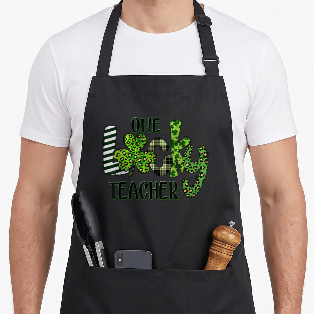 Shamrock teacher apron - One Lucky Teacher text with striped, plaid, leopard, and shamrock patterned letters in green — shown on a black cotton canvas apron against a white background