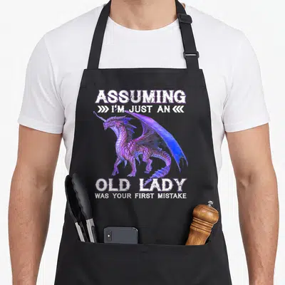 Assuming dragon apron - Realistic purple and blue dragon surrounded by white text assuming old lady mistake — shown on a black cotton canvas apron against a white background