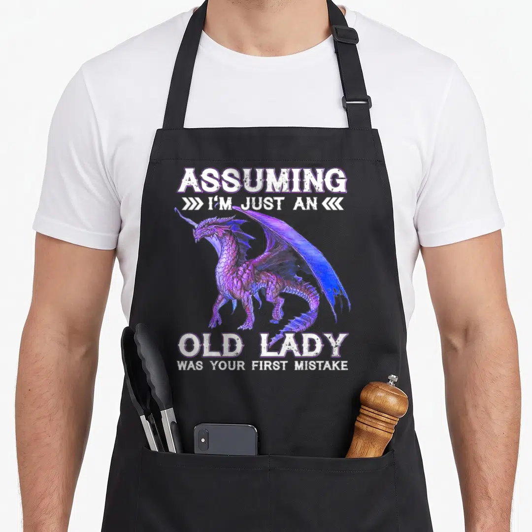 Assuming dragon apron - Realistic purple and blue dragon surrounded by white text assuming old lady mistake — shown on a black cotton canvas apron against a white background