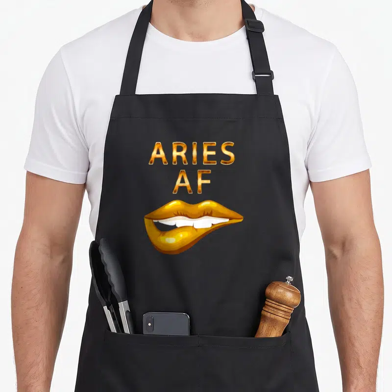Lip aries kitchen apron - Gold ARIES AF text above a pair of slightly parted lips showing white teeth — shown on a black cotton canvas apron against a white background