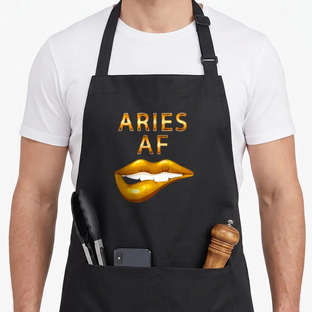 Lip aries kitchen apron - Gold ARIES AF text above a pair of slightly parted lips showing white teeth — shown on a black cotton canvas apron against a white background