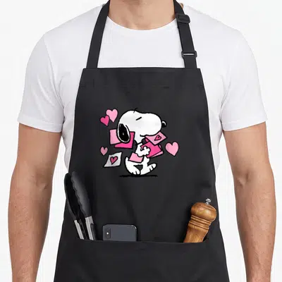 Letter valentine's day apron - Cartoon Snoopy holding pink cards with scattered pink hearts on a black background — shown on a black cotton canvas apron against a white background