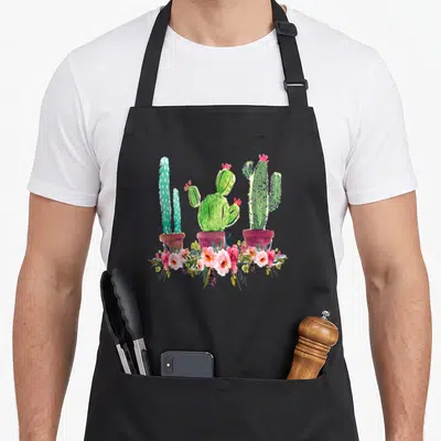 Blooming cactus apron - Three green potted cacti with pink and red flowers running along the bottom in watercolor style — shown on a black cotton canvas apron against a white background