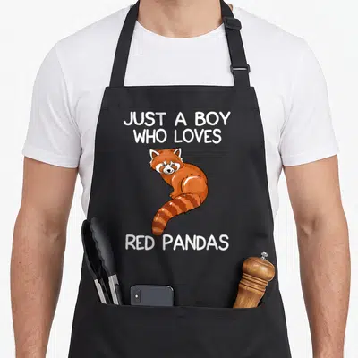 Vintage pandas panda apron - Cartoon red panda in orange and brown sandwiched between white text reading JUST A BOY WHO — shown on a black cotton canvas apron against a white background