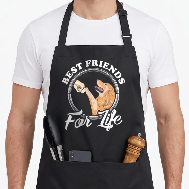 Funny golden retriever apron - Cartoon orange dog and human hand fist bumping inside white circle with best friends — shown on a black cotton canvas apron against a white background