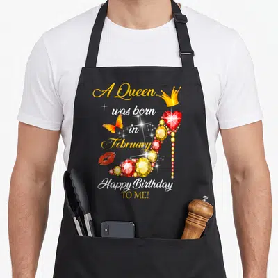 Vintage february birthday apron - Jeweled high heel with crown, butterfly, and red lips surrounding A Queen was born — shown on a black cotton canvas apron against a white background
