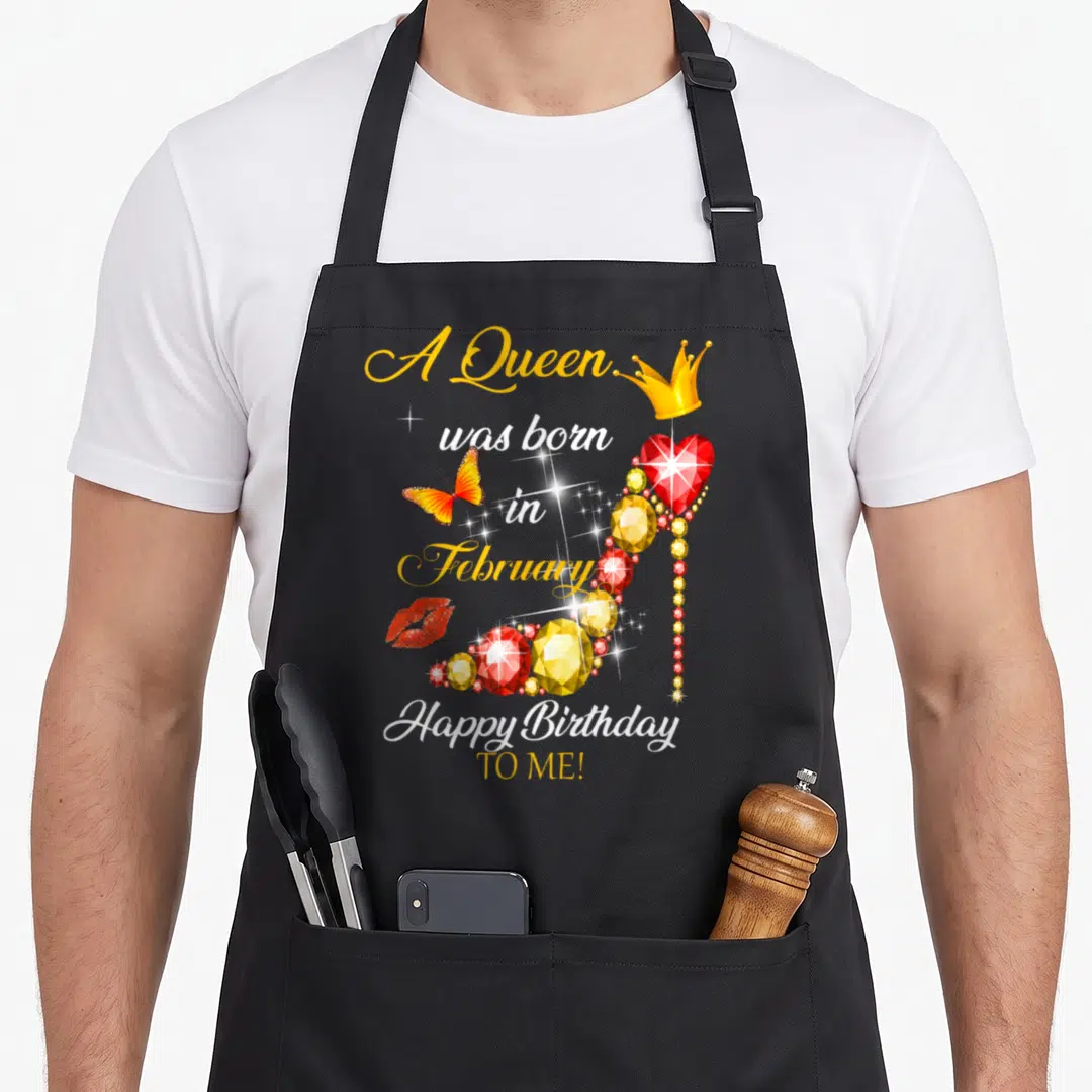 Vintage february birthday apron - Jeweled high heel with crown, butterfly, and red lips surrounding A Queen was born — shown on a black cotton canvas apron against a white background