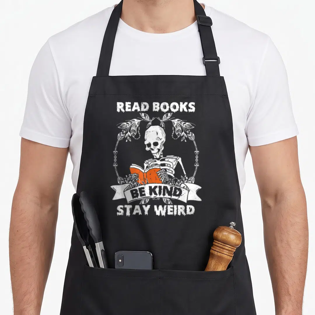 Read skeleton apron - White skeleton holding orange book inside leaf wreath with white text banners — shown on a black cotton canvas apron against a white background