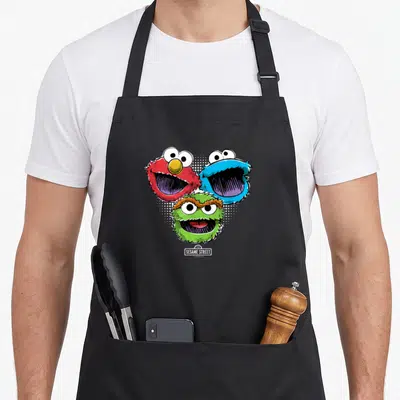 Halftone street apron - Three cartoon monster heads in red blue and green arranged in a triangle above a sesame street — shown on a black cotton canvas apron against a white background