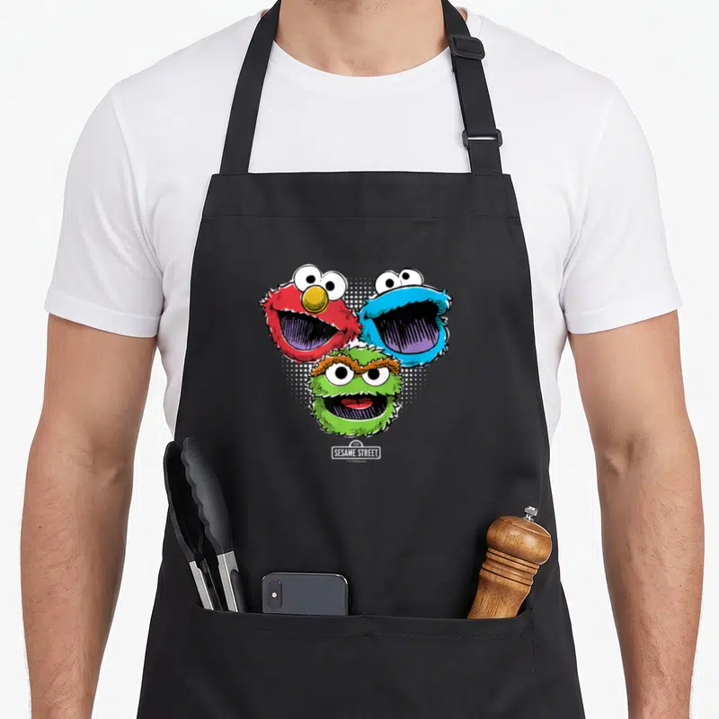 Halftone street apron - Three cartoon monster heads in red blue and green arranged in a triangle above a sesame street — shown on a black cotton canvas apron against a white background