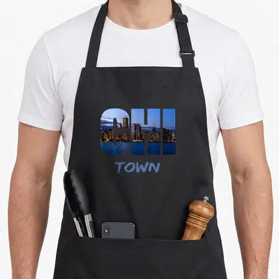 Chi windy chicago apron - CHI letters filled with city skyline photo above blue TOWN text — shown on a black cotton canvas apron against a white background