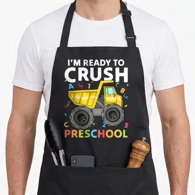 Preschool pre truck apron - Yellow dump truck with I'M READY TO CRUSH PRESCHOOL text and scattered colorful letters — shown on a black cotton canvas apron against a white background