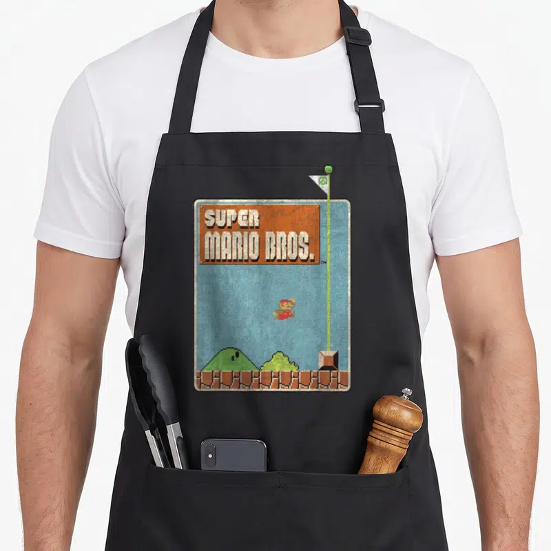 Bro game aprons - Pixel art Mario jumping near flagpole with SUPER MARIO BROS title and brick ground — shown on a black cotton canvas apron against a white background