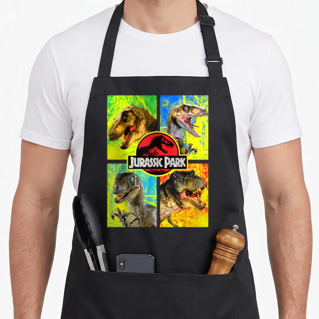 Four dinosaur apron - Four dinosaur heads in a grid with a central Jurassic Park logo in yellow, red, blue, and green — shown on a black cotton canvas apron against a white background