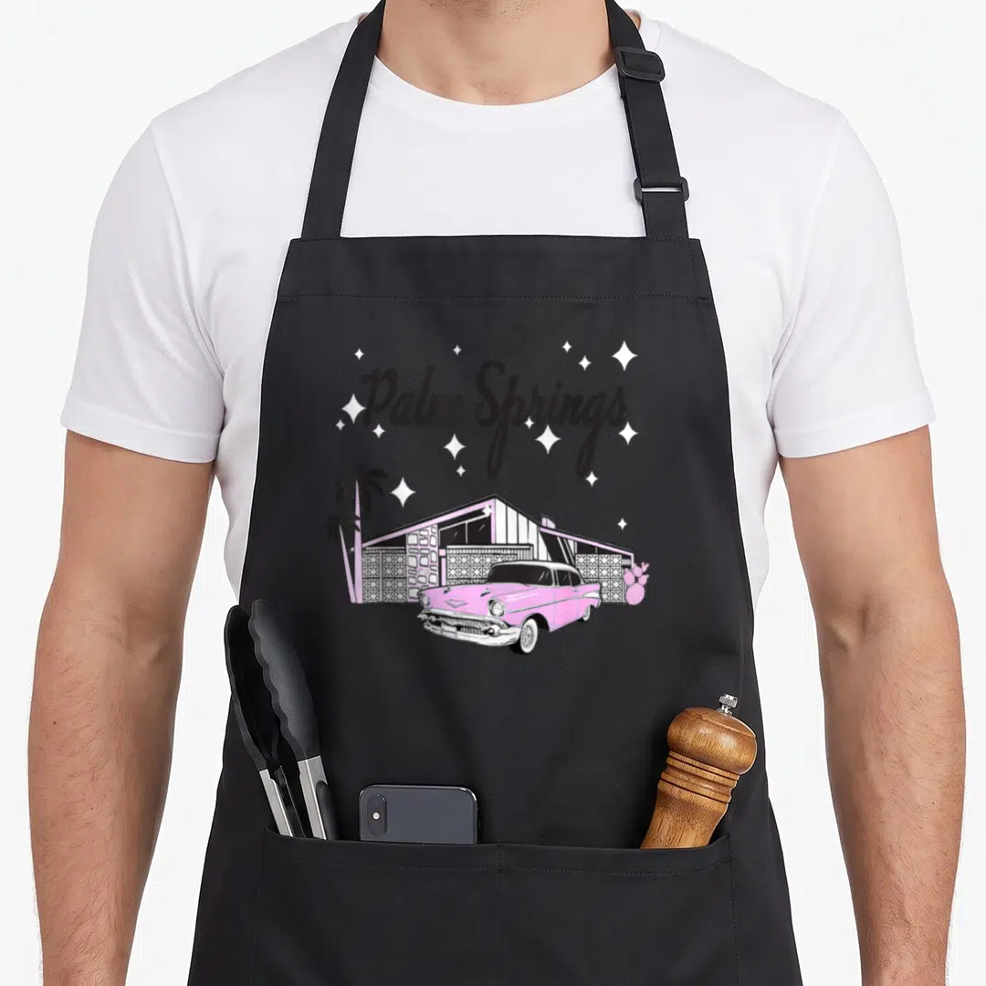 Mid spring apron - Palm Springs text above pink mid-century house, car, palm tree, and cactus with scattered stars — shown on a black cotton canvas apron against a white background