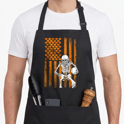 Fan halloween apron - skeleton in helmet holding football against orange flag with pumpkin stars — shown on a black cotton canvas apron against a white background