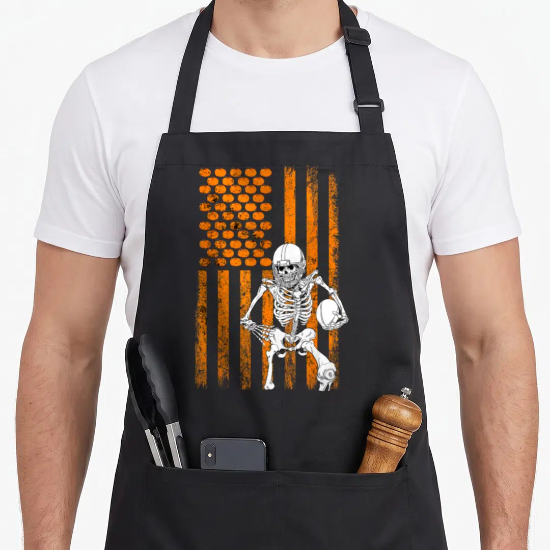 Fan halloween apron - skeleton in helmet holding football against orange flag with pumpkin stars — shown on a black cotton canvas apron against a white background