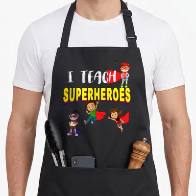 Teach kindergarten teacher apron - I TEACH SUPERHEROES text with three cartoon superhero children in yellow, red, blue — shown on a black cotton canvas apron against a white background