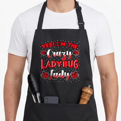 Collector ladybug apron - Stacked red text Yes I'm The Crazy Ladybug Lady flanked by four red ladybugs and scattered — shown on a black cotton canvas apron against a white background
