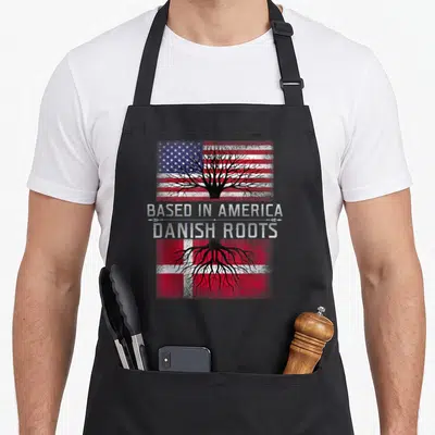 Danish pride apron - Black tree canopy over USA flag, roots over Denmark flag, stacked text — shown on a black cotton canvas apron against a white background