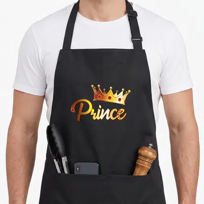 Prince family aprons - Gold crown and Prince script with red gems on black fabric — shown on a black cotton canvas apron against a white background