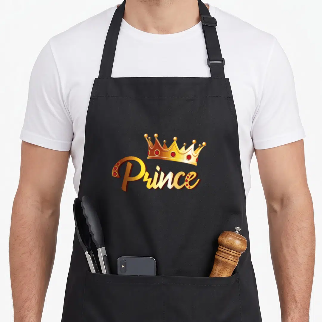 Prince family aprons - Gold crown and Prince script with red gems on black fabric — shown on a black cotton canvas apron against a white background