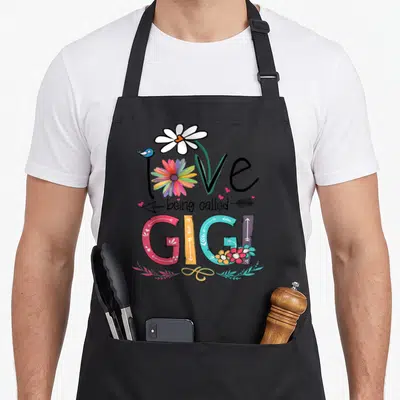 Gigi sunflower apron - Love being called Gigi text with rainbow flower, daisy, bird, and arrows in cartoon style — shown on a black cotton canvas apron against a white background