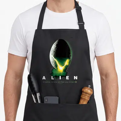 Space aliens apron - Cracked egg with green smoke and text ALIEN in space no one can hear you scream — shown on a black cotton canvas apron against a white background