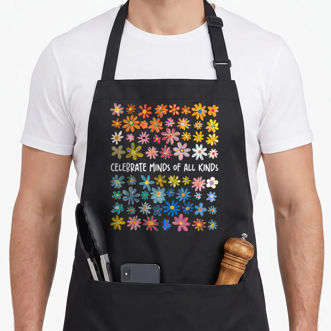 Mind celebrate diversity apron - Cartoon flowers in orange pink and blue surrounding centered white text CELEBRATE — shown on a black cotton canvas apron against a white background