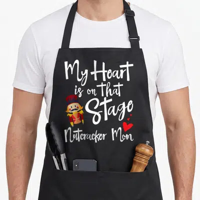 Ballet nutcracker apron - My Heart is on that Stage Nutcracker Mom text with cartoon nutcracker and three red hearts — shown on a black cotton canvas apron against a white background