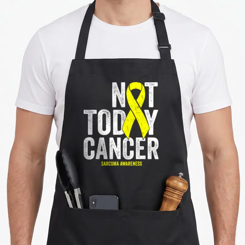 Today ribbon apron - Yellow ribbon replacing O in Not Today Cancer Sarcoma Awareness text — shown on a black cotton canvas apron against a white background