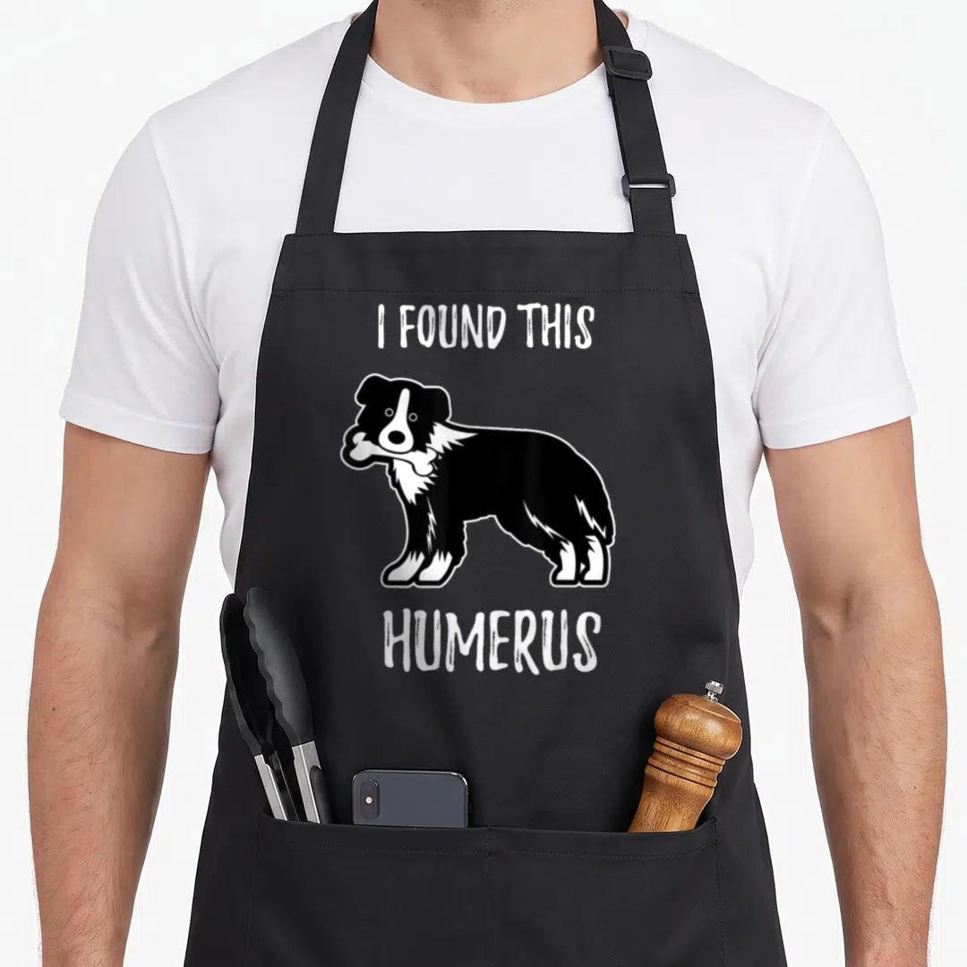 Dog pet border collie apron - Cartoon black and white dog holding a bone with text I FOUND THIS HUMERUS — shown on a black cotton canvas apron against a white background