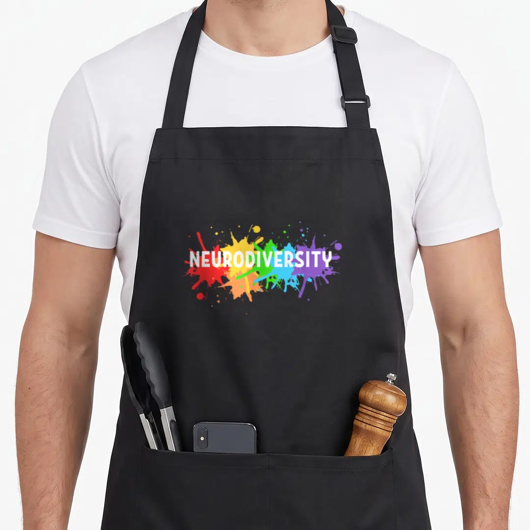 Neurodivergent adhd rainbow apron - White NEURODIVERSITY text over red orange yellow green blue purple paint splatters — shown on a black cotton canvas apron against a white background