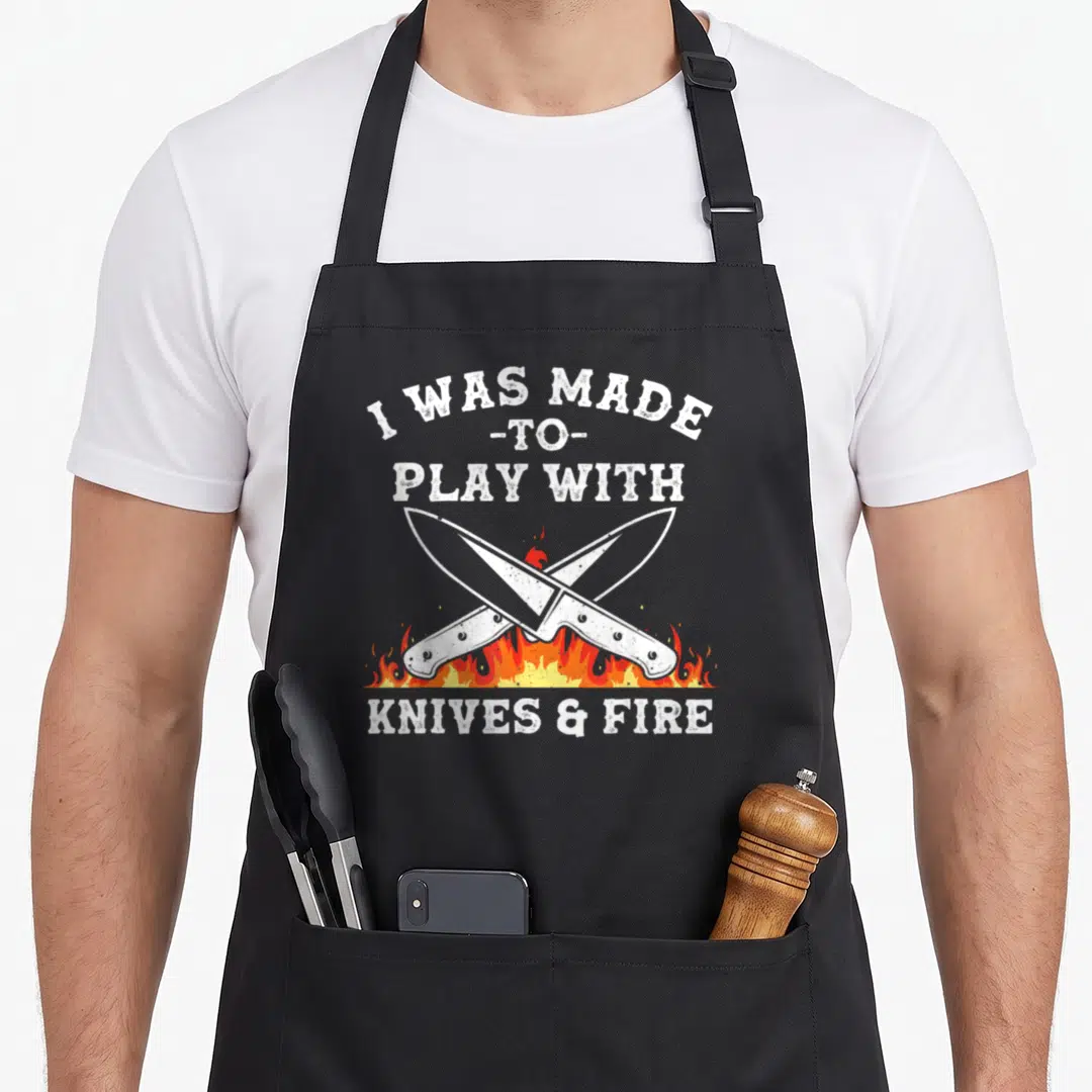 Knive cute culinary apron - Crossed knives over red orange flames with distressed white text reading I WAS MADE TO PLAY — shown on a black cotton canvas apron against a white background
