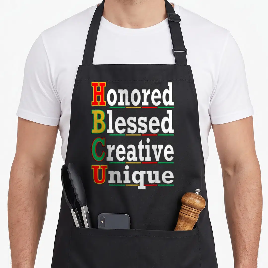 Honored creative apron - Stacked white text with red yellow green patterned initials and segmented underlines — shown on a black cotton canvas apron against a white background