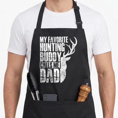 Hunting animal apron - White distressed text My Favorite Hunting Buddy Calls Me Dad next to deer head profile — shown on a black cotton canvas apron against a white background