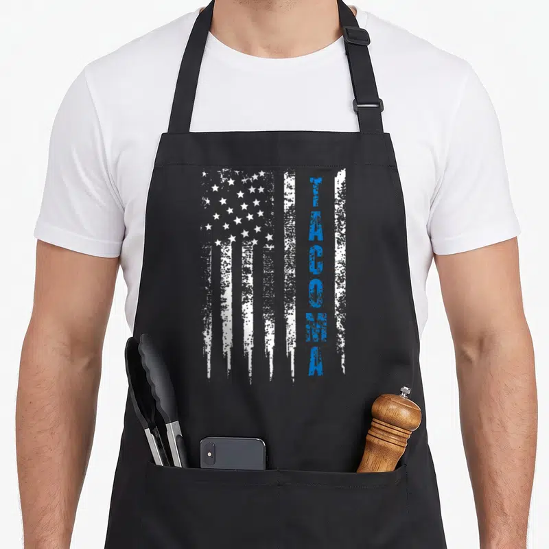 Tacoma line apron - Vertical distressed white flag stripes with blue Tacoma text overlaid — shown on a black cotton canvas apron against a white background