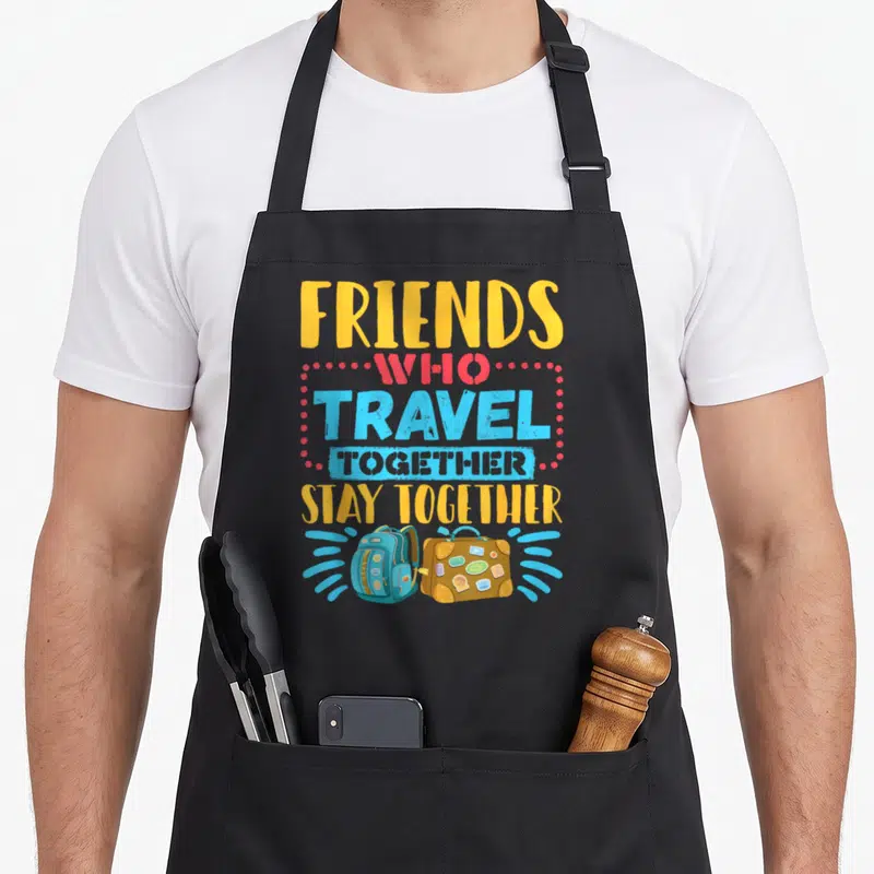Travel friends apron - Stacked text FRIENDS WHO TRAVEL TOGETHER STAY TOGETHER above cartoon blue backpack and brown — shown on a black cotton canvas apron against a white background