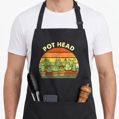Pot gardener apron - POT HEAD text over retro striped sun with row of green cacti in terracotta pots — shown on a black cotton canvas apron against a white background