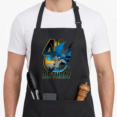 Poster birthday apron - Leaping Batman graphic with 4TH BIRTHDAY text in blue and yellow cartoon style — shown on a black cotton canvas apron against a white background