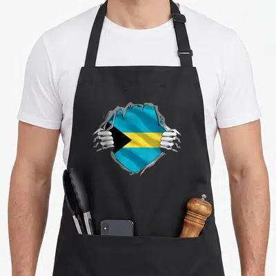 Independence bahamian apron - Two hands ripping grey surface to reveal blue yellow and black flag — shown on a black cotton canvas apron against a white background