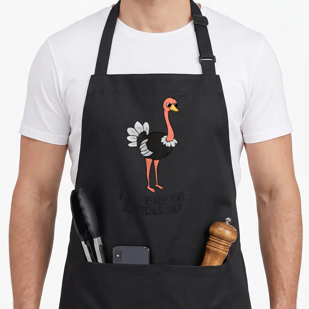 Vintage i love my ostrich apron - Cartoon ostrich with pink neck and text I JUST REALLY LIKE, OSTRICHES, OK? — shown on a black cotton canvas apron against a white background